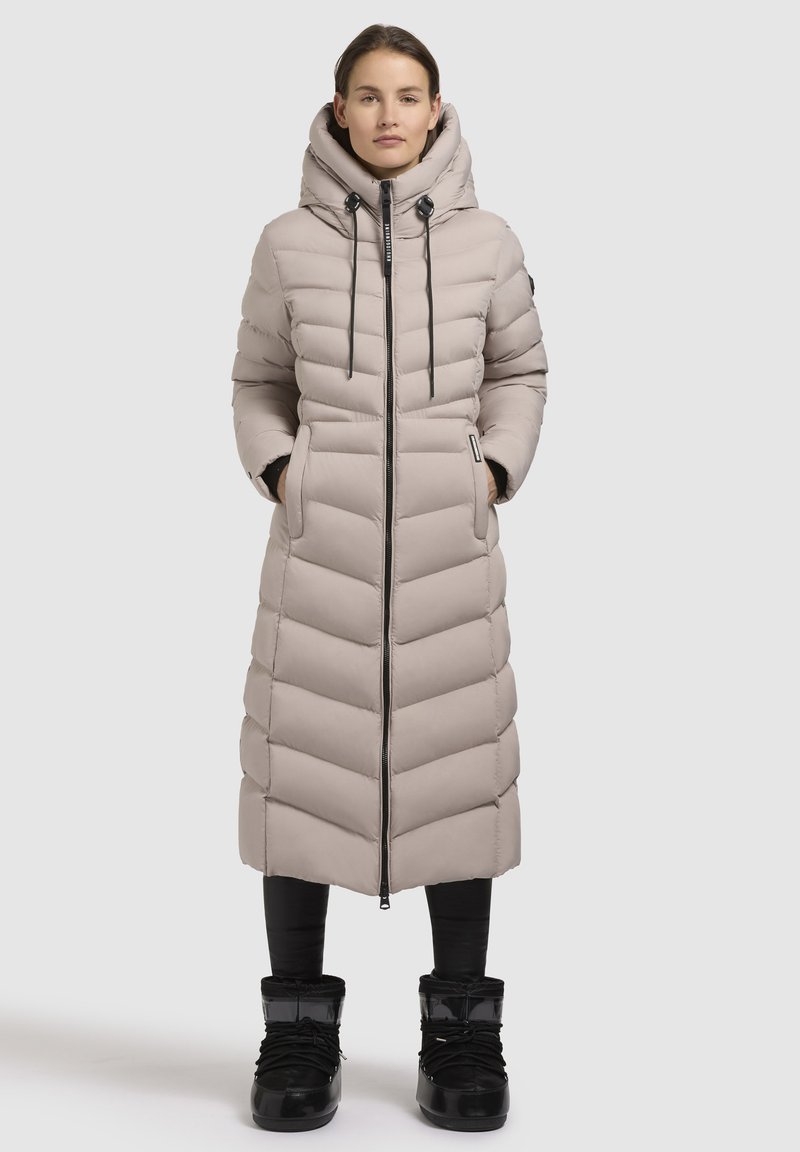 Bežinis ilgas pūkinis paltas su aukštu apykakle, zigzago formos siuvinėjimu, priekiniais užtrauktukais ir kapučonu su tamprėmis. Nešiojamas su juodomis legginsomis ir batais.