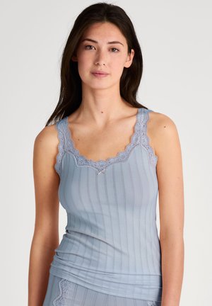 Jonge vrouw die een lichtblauw geribd hemd met kant afwerking en bijpassende onderkleding draagt, staand tegen een effen witte achtergrond.