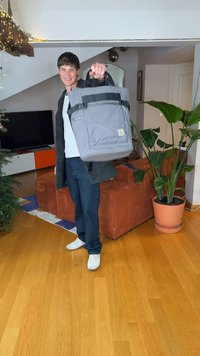 Man smiling indoors, holding a gray backpack extended toward the camera, standing near a couch, TV, and potted plant in a living room.
