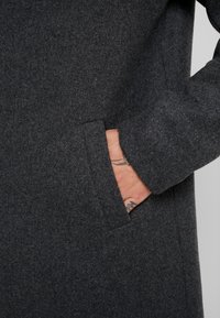 Cappotto di lana grigio scuro con una texture morbida, caratterizzato da una tasca laterale e una mano visibile che riposa all'interno della tasca.