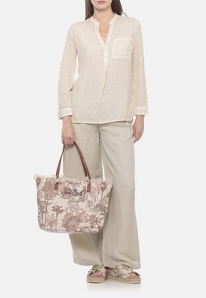 Donna con camicetta beige a maniche lunghe e pantaloni a gamba larga che tiene una borsa a mano beige con manici marroni e fantasia.