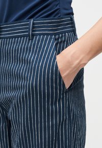 Main reposant dans la poche d'un pantalon bleu à rayures verticales blanches avec une ceinture rayée assortie.