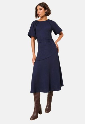 Marineblaue Midikleid mit kurzen, weiten Ärmeln, strukturiertem Stoff, geschwungener Saumlinie, kombiniert mit braunen kniehohen Stiefeln mit Absatz.