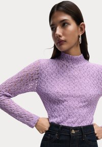 Top lungo a maniche con pizzo viola, scollatura alta, motivo floreale intricato e silhouette aderente, abbinato a jeans scuri a vita alta.