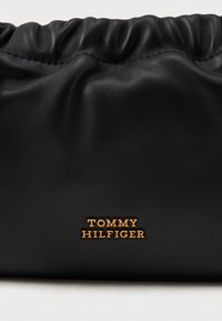 Černá kožená taška s nabíraným vrškem. Obsahuje zlaté logo "TOMMY HILFIGER". Hladký povrch, střední velikost, vhodná pro každodenní použití.