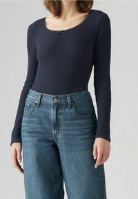 Kvinna som bär en marinblå ribbad långärmad topp med rundad halsringning och högmidjade mellanblå jeans.