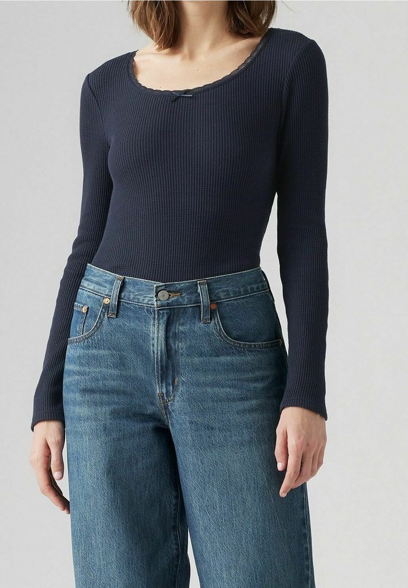 Kvinna som bär en marinblå ribbad långärmad topp med rundad halsringning och högmidjade mellanblå jeans.