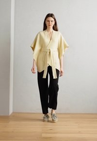 Haut en laine jaune avec des manches larges et un lien à l'avant, associé à un pantalon noir et des chaussures grises. Texture lisse, design simple.