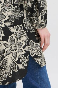 Blusa nera floreale con fiori color crema dai dettagli intricati e una texture stropicciata. Include una fessura laterale e polsini elasticizzati.