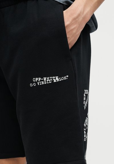 Gros plan d'une personne portant un short noir avec le texte blanc Off-White c/o Virgil Abloh™, la main dans la poche avant, sur un fond uni.