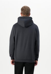 Man wearing a plain dark gray hoodie and black pants, standing facing away against a white background.