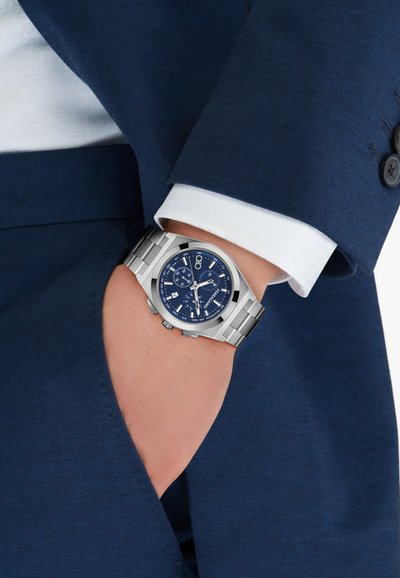 Srebrni kronografski sat s plavim brojčanikom, metalnom narukvicom i teksturiranim licem. Nošen na zapešću s plavim odijelom i vidljivim bijelim manšetama košulje.