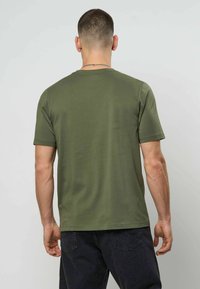 Mann med kort hår iført en olivengrønn kortermet t-skjorte og mørke jeans, stående med ryggen mot kameraet.