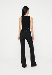 Woman with long black hair wearing a sleeveless black top with zipper and fitted black pants, standing against a plain white background.