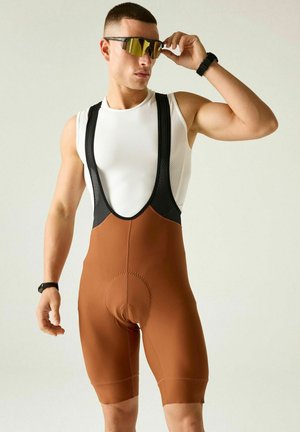 Atleta con culottes de ciclismo marrones con tirantes, camiseta interior sin mangas blanca, ajustándose gafas de sol deportivas reflectantes, de pie frente a un fondo claro y liso.
