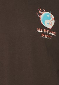 T-shirt in cotone marrone con grafica yin-yang blu e bianca decorata da fiamme rosse e la scritta "TUTTO CIÒ CHE HAI È ADESSO" in bianco e rosso.