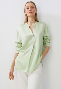 Blusa verde chiaro con un design senza colletto, maniche lunghe e una vestibilità rilassata. Texture liscia, leggera lucentezza e tasche laterali.