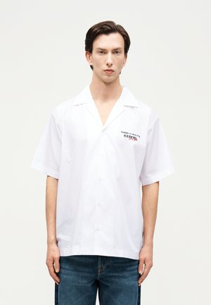 Νεαρός με σκούρα μαλλιά φοράει λευκό πουκάμισο με κοντά μανίκια και κουμπιά, με ένα μικρό λογότυπο στο αριστερό στήθος και μπλε τζιν.