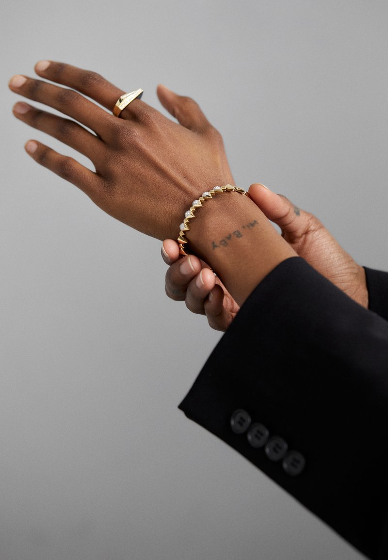 Zelta tonis rotaslietas ietver teksturētu gredzenu un aproci ar mainīgām trīsstūra un apaļām formām, kas valkā uz tumšas ādas roku melnā piedurknē.