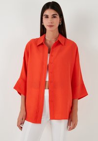 Red oversized button-up shirt with a collar and wide sleeves, made of a lightweight fabric, paired with a white cropped top and pants.