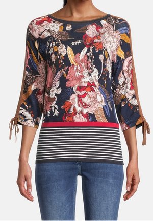 Kvinde iført en navyblå blomsterprintet top med brune båndærmer og en rød- og sortstribet kant, kombineret med blå jeans.
