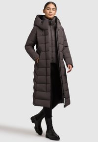 Frau trägt einen langen dunkelbraunen gepolsterten Mantel mit großer Kapuze, schwarze Leggings und schwarze klobige Stiefeletten, steht mit den Händen in den Taschen.