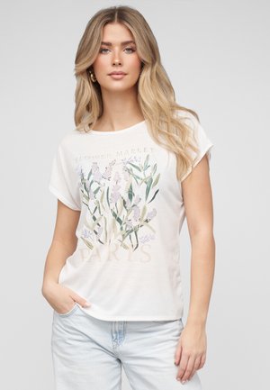 Giovane donna con lunghi capelli biondi che indossa una maglietta bianca con stampa floreale e jeans azzurri, in piedi con una mano in tasca.