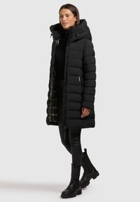 Svart pufferjacka med en bred huva, dragkedja fram och ribbade ärmar. Vadderad struktur, lång längd och slimmad design. Kombinerad med svarta läderbyxor och stövlar.