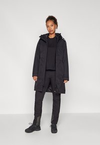 Sort vandtæt parka-jakke med hætte, med lynlås-lukning, to sidelommer og elastiske manchetter. Båret over sorte basislag og bukser.