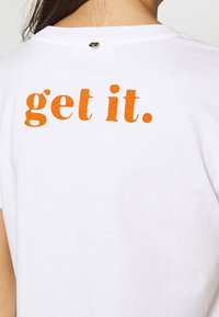 Osoba v bílém tričku s velkým oranžovým textem "get it." vytištěným na horní části zad pod výstřihem.