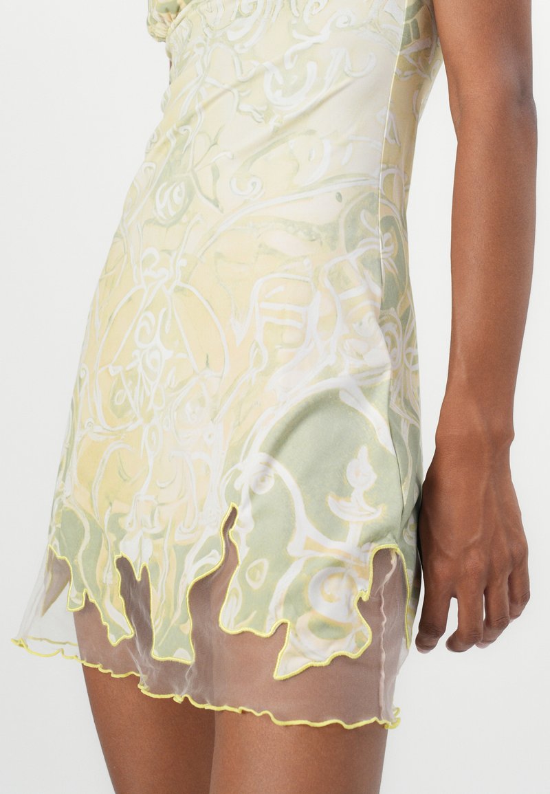 Przezroczysta mini sukienka w jasnym żółtym kolorze z zielonymi, misternymi spiralnymi wzorami, z falbankowym dołem obszytym jasnym żółtym ściegiem.