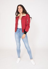 Veste bomber rouge avec un col en fausse fourrure crème, poche zippée sur la manche gauche et poignets côtelés ; associée à un jean skinny bleu clair.