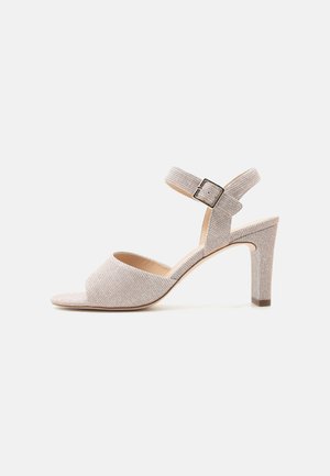 Sandale à talon haut ouverte beige avec bride à la cheville et boucle argentée, tissu texturé, et talon bloc sur fond blanc.