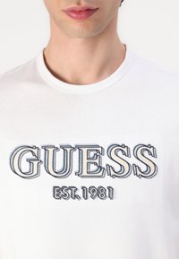 Weißes Baumwoll-T-Shirt mit einem großen bestickten "GUESS"-Logo in Blau und Grau mit "EST. 1981" darunter. Klassischer Rundhalsausschnitt.