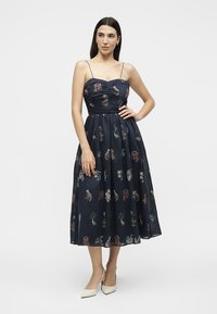 Femme portant une robe midi bleu marine avec de fines bretelles et des motifs brodés multicolores, associée à des escarpins blancs à bout pointu.