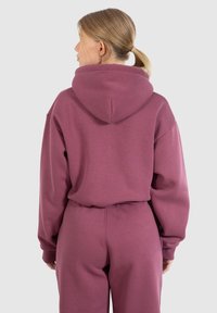 Felpa con cappuccio oversize di colore rosa, con una texture morbida e tasca a marsupio, abbinata a pantaloni della tuta coordinati, vista da dietro.