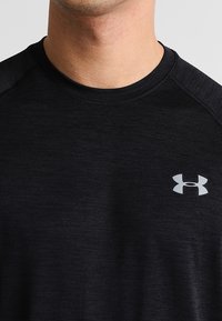 Černé atletické tričko s kulatým výstřihem, krátkými rukávy a jemnou melírovanou texturou. Na hrudi je malý stříbrný logo.