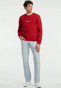 Jauns vīrietis, kas skatās pa kreisi, uzvilcis sarkanu Guess Jeans džemperi, gaiši zilas džinsus un baltas kedas, stāvot pret vienkāršu gaismas fonu.