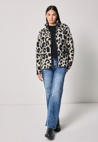 Chaqueta de leopardo en piel sintética de color crema y negro, con cierre de cremallera en la parte delantera, dos bolsillos laterales, combinada con vaqueros azules y botines negros.
