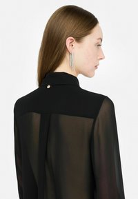 Blusa nera trasparente con colletto alto, dotata di un piccolo bottone dorato sul retro. La modella indossa orecchini pendenti argentati scintillanti.