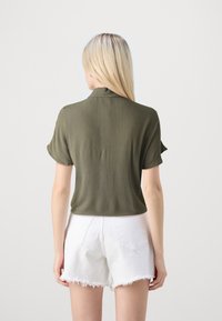 Top corto a maniche corte verde oliva con vestibilità rilassata e colletto strutturato, abbinato a pantaloncini di jeans bianchi con orlo sfrangiato. Tessuto complessivamente morbido.
