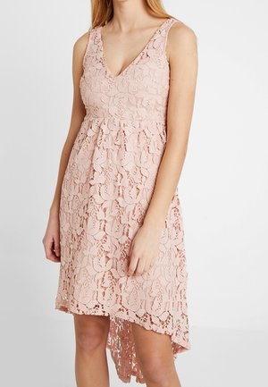 Robe rose en dentelle sans manches avec motif floral, encolure en V, taille cintrée et ourlet asymétrique, portée par une personne debout.