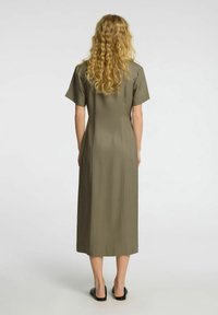 Oliven grønn midikjole i glatt stoff. Korte ermer og rett snitt med subtile sidespalter. Brukt med svarte slip-on sko.