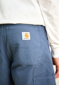 Blaue Arbeitshose aus Canvas mit mehreren Taschen und einem Carhartt-Logo-Patch in Gelb und Orange auf der Gesäßtasche.