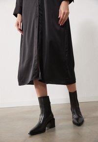 Abito lungo in satin nero con chiusura a bottoni e spacco laterale; abbinato a stivali neri in pelle liscia con punta quadrata e tacco a blocco.