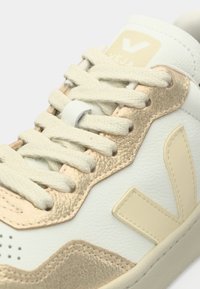 Άσπρο και μπεζ παπούτσι Veja με κρέμ κορδόνια και μεταλλικές μπεζ λεπτομέρειες, που δείχνει υφή δέρματος και ραμμένη λεπτομέρεια λογότυπου.