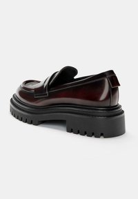 Dark brown leather loafer with thick black chunky sole and a strap across the upper, photographed against a plain white background.