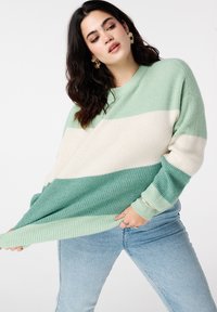 Pull rayé vert avec une coupe décontractée, une texture côtelée et des blocs de couleur vert clair, vert foncé et crème. Porté avec un jean bleu clair.