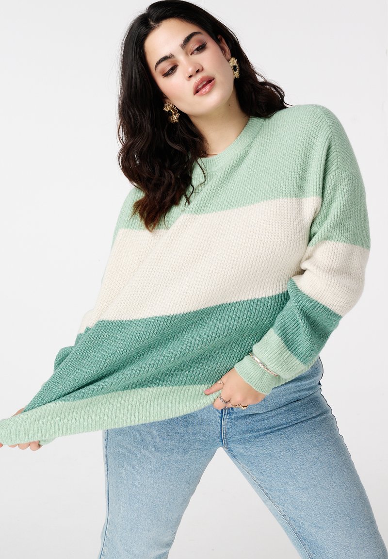 Pull rayé vert avec une coupe décontractée, une texture côtelée et des blocs de couleur vert clair, vert foncé et crème. Porté avec un jean bleu clair.