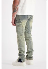 Lichtblauwe denim cargo pants met meerdere zakken, tapered fit, vervaagde textuur en ritsen bij de enkel, gecombineerd met witte sneakers.